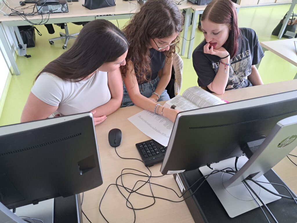 Drei Schülerinnen schauen gemeinsam auf ein Dokument und einen Computerbildschirm in einem Klassenraum.