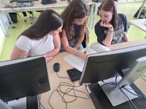 Drei Schülerinnen schauen gemeinsam auf ein Dokument und einen Computerbildschirm in einem Klassenraum.