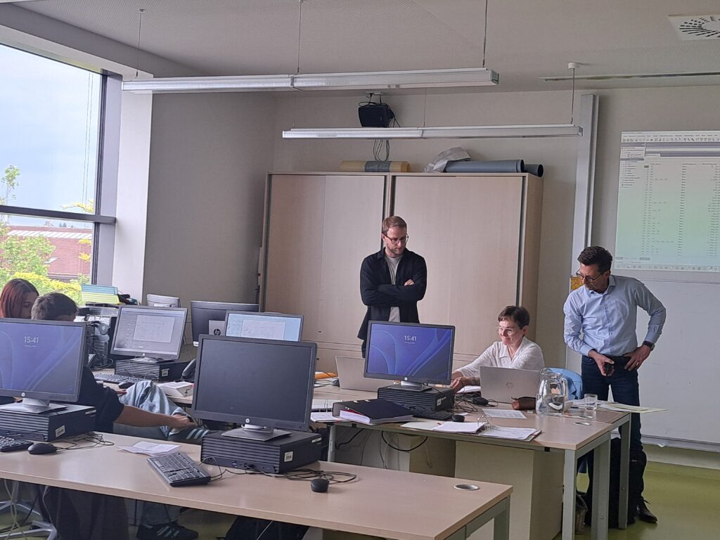 Drei Personen stehen in einem Schulungsraum, während andere an Computern arbeiten.