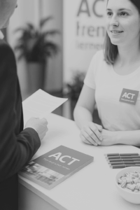 Mitarbeiterin am Empfangstresen mit Informationsmaterial und einem Besucher im Gespräch.